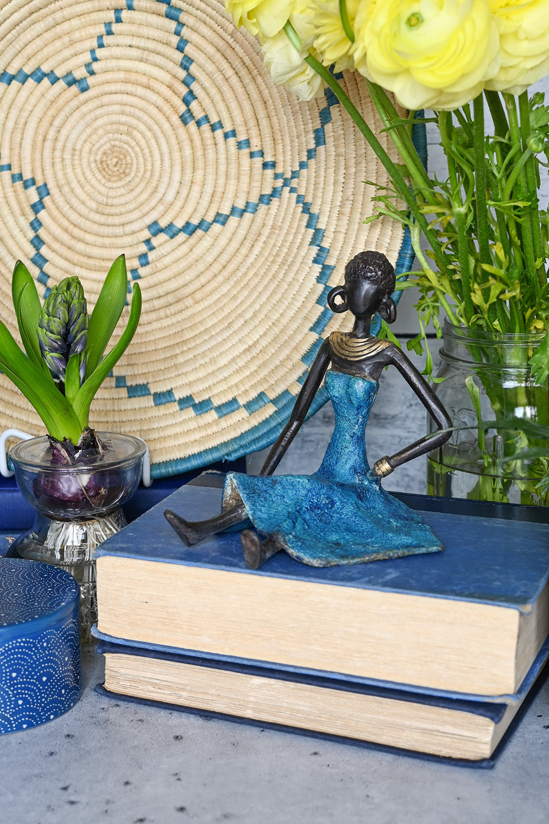 Bronze sculpture from Burkina Faso | statue of seated woman wearing blue dress and large hoop earrings, posed on top of old books